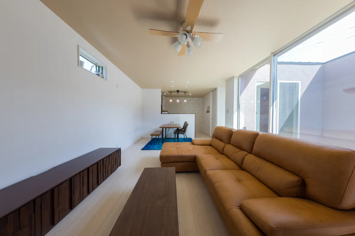 広い窓から自然光が差し込む明るいリビング、ブラウンのレザーソファと木製ローテーブルが配置された空間