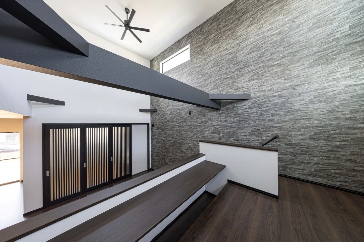 吹き抜け空間に大胆に渡された黒い梁と石調アクセントウォールが印象的な、モダンで開放的なリビング上部の様子