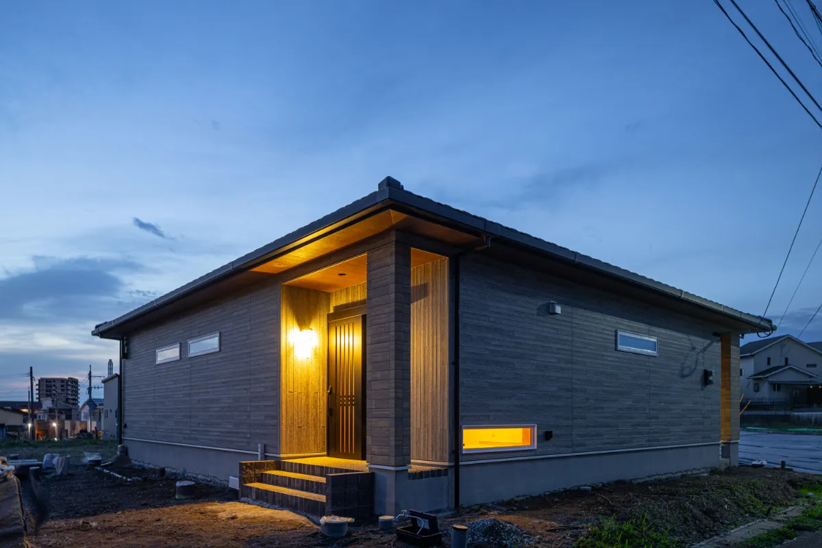玄関ポーチにあたたかな灯りがともる夕暮れの外観。木目の軒天と落ち着いた外壁が、やさしい雰囲気をつくっています。