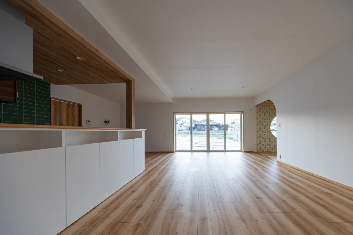 キッチン横から見たリビング全体。木目の床が奥まで続き、大きな掃き出し窓から自然光が入る開放的な空間です。