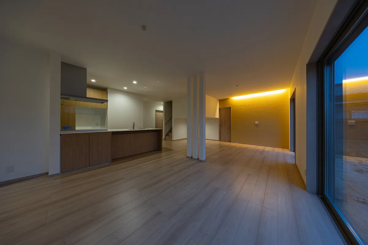 夜のリビング空間。壁面の間接照明がアクセントとなり、木目の床とキッチンの素材感が引き立つ、あたたかみのある室内風景です。