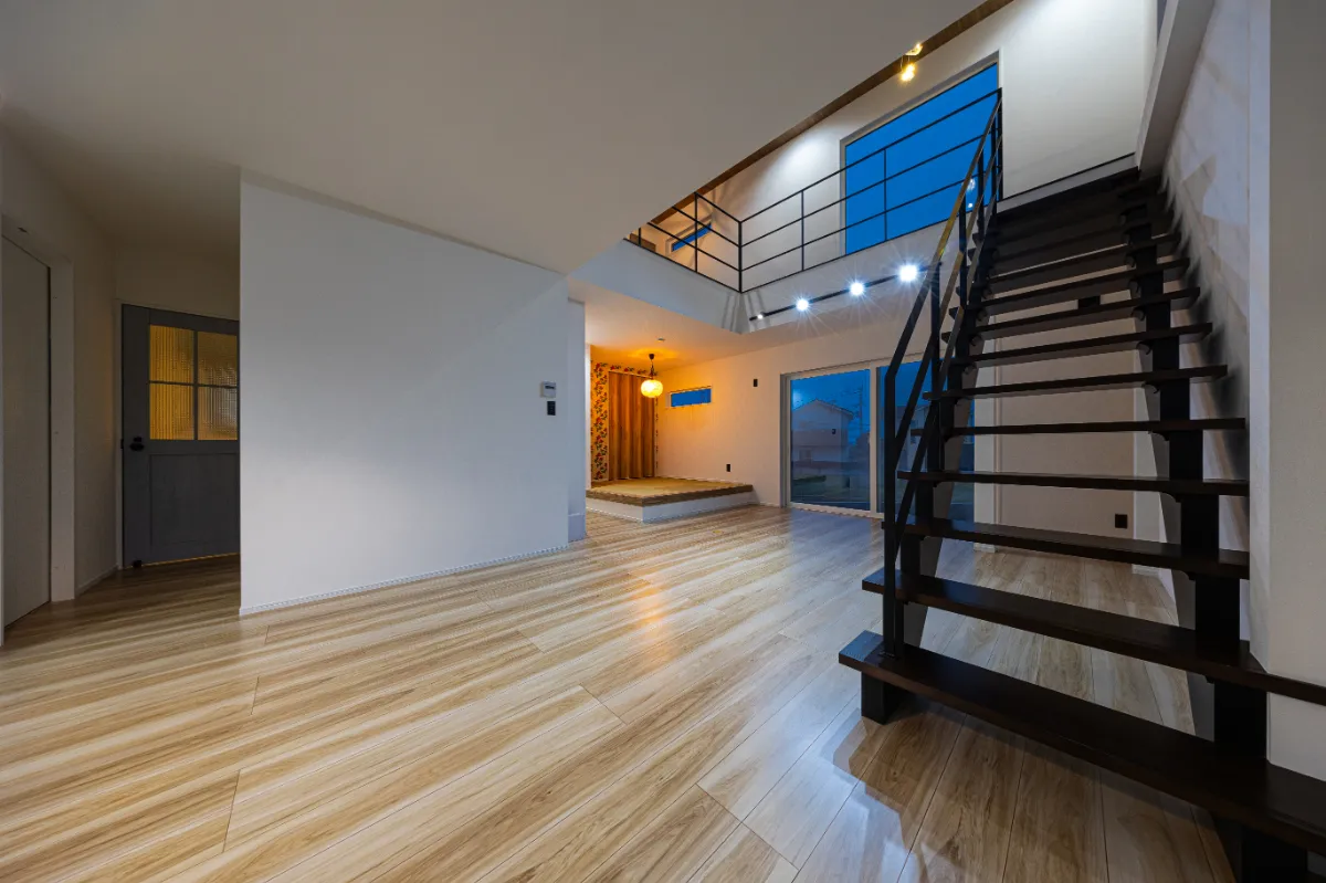 小上がりスペースと吹き抜けを望む夜のリビング。階段まわりの照明が空間に立体感を与え、家族のくつろぎ時間を包み込みます。