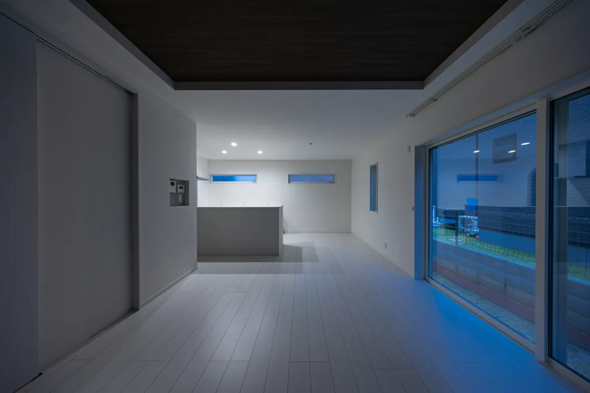 夜のリビング全体。白い床に落ち着いた折り上げ天井を合わせ、キッチンまで視線が抜ける、開放感のある空間です。