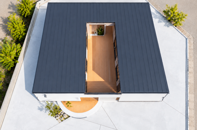 上空から撮影した、中庭のあるモダンな住宅の外観。ダークグレーの屋根に囲まれた細長いウッドデッキが特徴的で、白いコンクリートの敷地と周囲の緑が調和した、スタイリッシュなコの字型の住まい。